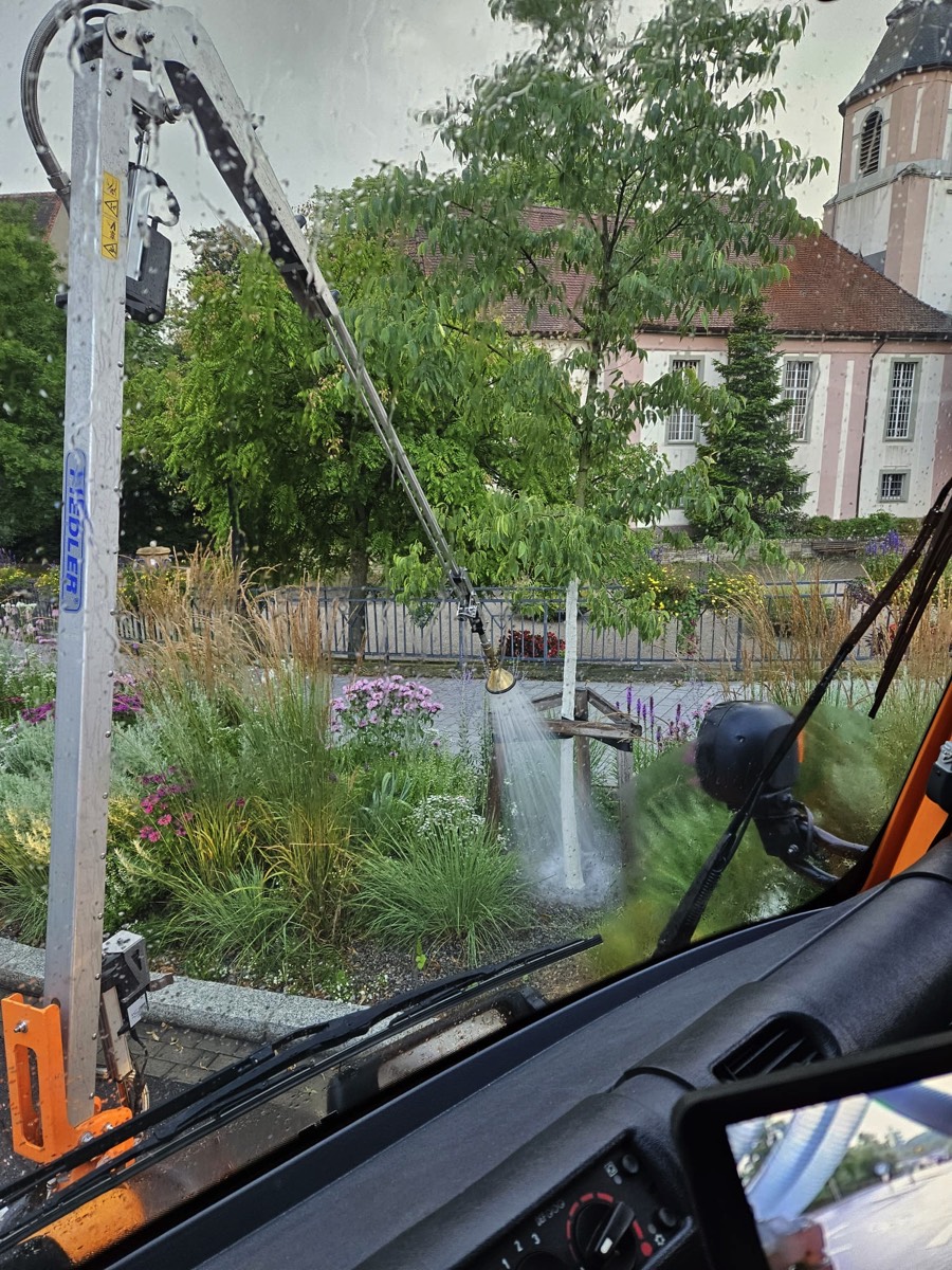 Ein mechanischer Arm mit einem Schlauch führt eine Bewässerung durch und bewässert einen neu gepflanzten Baum, der von Holzpfählen gestützt wird und sich in einer grasbewachsenen Grünfläche in Baden-Württemberg befindet, die von weiteren Bäumen und üppiger Vegetation umgeben ist.